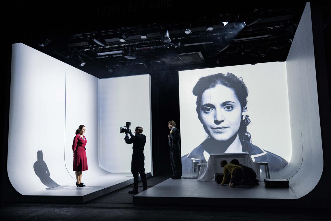A person in a red dress standing in front of person with a camera in a white room while they have their face projected in black and white onto one of the walls.